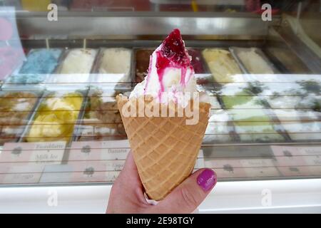 Una mano di donna che tiene un unico cono gelato riempito con rinomato gelato italiano condito con salsa di amarena, scelta da una varietà di Fla Foto Stock
