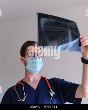 un bel medico che indossa scrub, stetoscopio e maschera facciale che guarda a raggi x durante la pandemia di sars cov 2 Foto Stock