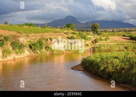 Sendrisoa, Madagascar - 28 aprile 2019: Fiume lento fangoso, erba verde e cespugli su entrambi i lati, piccole montagne sullo sfondo, donna lava i vestiti Foto Stock