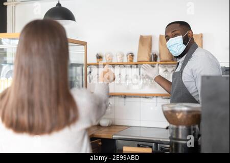 Un cameriere afroamericano maschio, un proprietario di caffè che indossa maschera e guanti si trova dietro il banco, una cliente donna fa un ordine. Un multirazziale Foto Stock