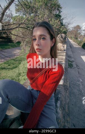 Immagine angolare di giovane donna pensierosa che guarda di fronte a. la fotocamera in una giornata invernale di sole mentre si è seduti Una panchina di fianco ad un Park.Seasonal Foto Stock