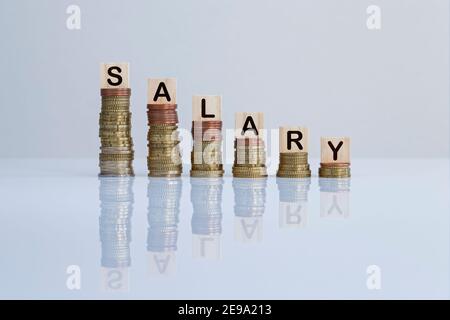 Parola 'SALARY' su blocchi di legno in cima a pile discendenti di monete in grigio. Foto concettuale di perdere denaro, crisi economica, recessione e fallimento. Foto Stock