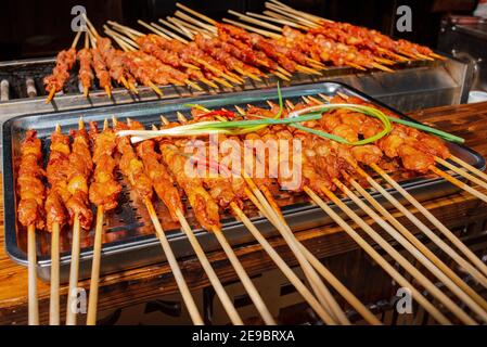 Cibo di strada. Cibo cinese. Succosi spiedini di pollo su spiedini in salsa piccante. Con pepe e cipolla. Sdraiati su un vassoio in metallo su un tavolo di legno. Primo piano. Foto Stock