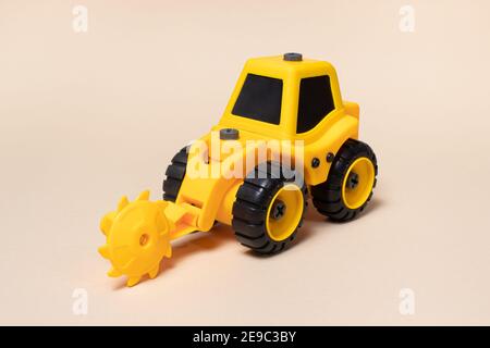 Trattore giallo giocattolo con ugello per sega circolare per legno su sfondo beige chiaro. Apparecchi per macchine da costruzione per il negozio di giocattoli. Foto Stock