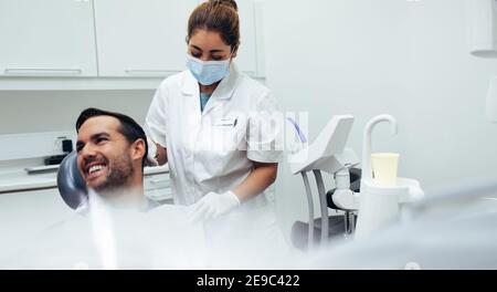 Paziente maschile seduto sulla sedia del dentista guardando via e sorridendo con un'infermiera femminile che mette sul grembiule dentale sul paziente. Dentista che assiste un paziente Foto Stock