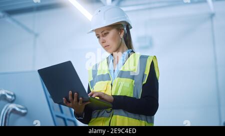 High-Tech Factory: Ingegnere femminile sicuro e professionale che indossa una giacca di sicurezza e un Hard Hat che tiene e lavora su un computer portatile. Moderno e luminoso Foto Stock
