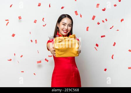 Felice donna asiatica sorridente in costume rosso tradizionale dando oro confezione regalo in grigio chiaro su sfondo studio con confetti per Concetti di anno nuovo cinese Foto Stock