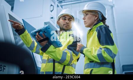Capo ingegnere e project manager che indossa giubbotti di sicurezza e copricapo rigidi, utilizza il controller digitale per tablet in fabbrica moderna, parlando, Programming Machine Foto Stock