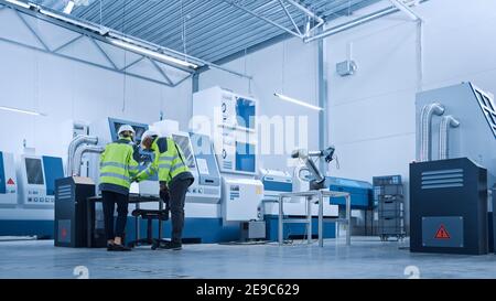 Capo ingegnere e project manager che indossa giubbotti di sicurezza e copricapo rigidi, utilizza il controller digitale per tablet in fabbrica, ottimizzando i macchinari CNC, programmazione Foto Stock