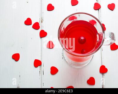 Fuoco selettivo sui mirtilli rossi in una bevanda fresca in una tazza di vetro su uno sfondo di legno bianco. Molti piccoli cuori rossi come simbolo del giorno di San Valentino Foto Stock
