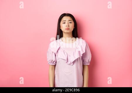 Triste pupazzare ragazza asiatica e accigliare, guardando sconvolto su qualcosa di ingiusto, lamentandosi, in piedi in vestito contro sfondo rosa Foto Stock