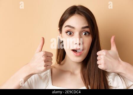 Molto fresco. Ragazza eccitata che mostra i pollici in su e gastping stupito, lodando il buon lavoro, complimento qualcosa di impressionante, in piedi su sfondo beige Foto Stock