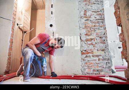 Vista orizzontale e laterale istantanea di idraulico bearded installazione di sistema di riscaldamento a pavimento in camera vuota con pareti in mattoni. Uomo che indossa tute e guanti, in piedi su un ginocchio, che lavora con un perforatore Foto Stock