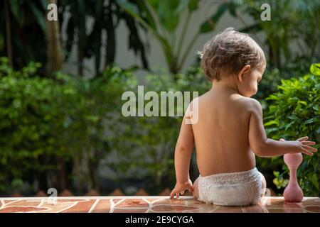 un affascinante bambino si siede con un giocattolo sui gradini di una casa sotto la luce del sole. vista posteriore. Foto Stock