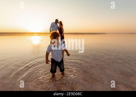 Ritratto di ragazzo di razza poco misto. Baciare i genitori sullo sfondo. Felice famiglia multietnica in spiaggia. Bellissimo tramonto Foto Stock