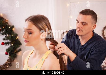Papà in pigiama intreccia la coda di sua figlia al mattino. Un uomo passa attraverso i capelli, fa i capelli della sua figlia giovane. Foto Stock