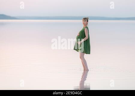 Elegante donna incinta nel vestito verde che posa al tramonto, guardando la macchina fotografica. Incredibile tramonto su un lago poco profondo. Foto Stock