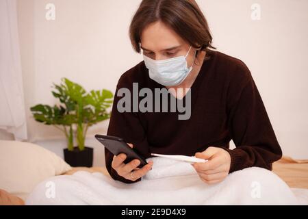 Un uomo malato chiama un'ambulanza al telefono. Un uomo mascherato a casa controlla la temperatura e compone il numero del medico sul telefono. Coronavirus, casa Foto Stock