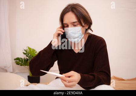 Un uomo malato chiama un'ambulanza al telefono. Un uomo mascherato a casa controlla la temperatura e compone il numero del medico sul telefono. Coronavirus, casa Foto Stock