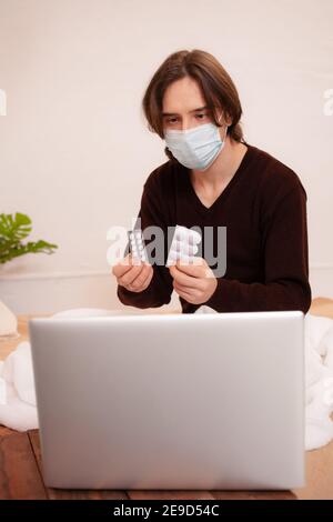 Un uomo malato si siede di fronte a un computer portatile e mostra al medico le sue pillole. Quarantena domestica, consultazione medica online. Un uomo che indossa una maschera a casa. Foto Stock