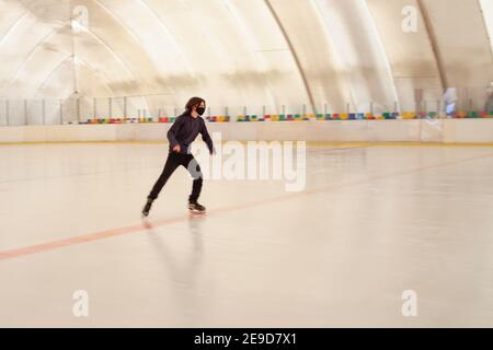 Un uomo mascherato pattina sul ghiaccio. Pista di pattinaggio interna. Sedili multicolore sullo sfondo Foto Stock