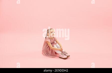 Seduto, sognando. Infanzia e sogno sul grande e famoso futuro. Bella ragazza longhair su sfondo corallo rosa studio. Infanzia, sogni, immaginazione, educazione, espressione facciale, concetto di emozioni. Foto Stock