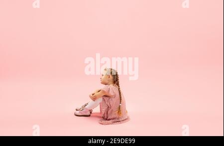 Seduto, sognando. Infanzia e sogno sul grande e famoso futuro. Bella ragazza longhair su sfondo corallo rosa studio. Infanzia, sogni, immaginazione, educazione, espressione facciale, concetto di emozioni. Foto Stock