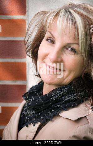 Primo piano laterale di una donna di mezza età che la appoggia contro una parete esterna e guarda la fotocamera con la testa inclinata. Foto Stock