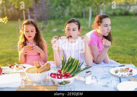 Albero bambina carina seduto al tavolo in natura e mangiare il pranzo. Divertirsi e parlare il giorno estivo soleggiato, Foto Stock