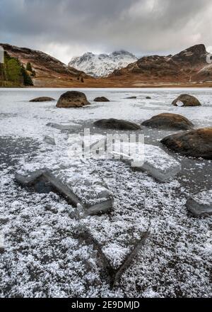 Ghiaccio incrinato coperto di fiocchi di neve con vista sulla montagna innevata con nuvole invernali scure nel cielo. Blea Tarn, Lake District, Regno Unito. Foto Stock