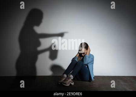 Violenza fisica domestica, abuso. Spaventata piccola ragazza caucasica, vittima seduta vicino alla parete bianca con l'ombra di arrabbiata minaccia madre con dipendenza da alcol. Consapevolezza del problema sociale. Foto Stock