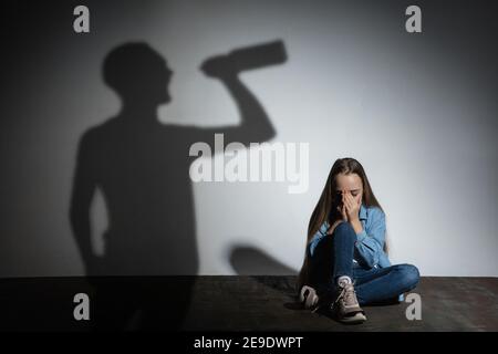 Violenza fisica domestica, abuso. Spaventata piccola ragazza caucasica, vittima seduta vicino alla parete bianca con ombra di arrabbiato minacciando padre con dipendenza da alcol. Consapevolezza del problema sociale. Foto Stock