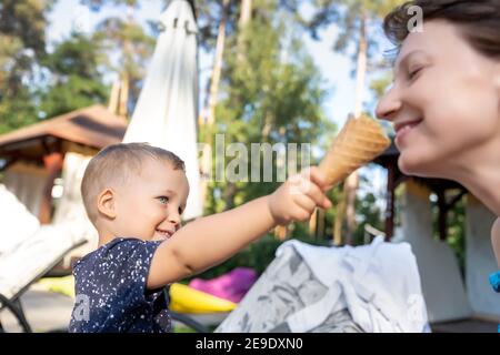 Piccolo adorabile e giocoso ragazzo biondo caucasico che condivide con la mamma il gelato dolce e gustoso durante la calda giornata estiva all'aperto. Due persone felici di godere Foto Stock