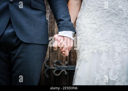 Primo piano di sposa in abito bianco e sposo in suit appena sposato tenendo le mani insieme dopo la cerimonia di nozze si è levato in piedi di fronte alla porta della chiesa in legno Foto Stock