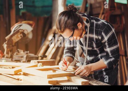 Un giovane falegname maschile che lavora nel complesso equivale ad una barra di legno con una fresatrice in officina, sullo sfondo in legno Foto Stock