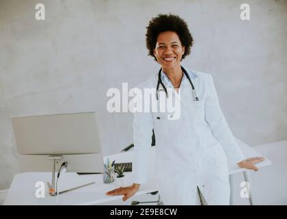 Donna Africana americana medico indossando camice bianco con stetoscopio in piedi dalla scrivania in ufficio Foto Stock