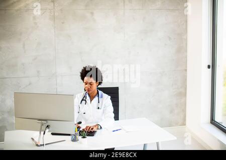 Donna Africana americana medico indossando camice bianco con stetoscopio seduta dietro la scrivania in ufficio Foto Stock