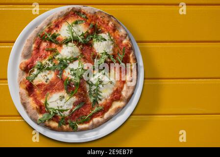 Pizza con salsa di pomodoro, mozzarella e rucola o razzo in piatto bianco su tavole gialle, piatto in vista dall'alto con spazio per la copia Foto Stock