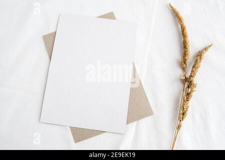 Scena mock-up di cancelleria in un tasto di luce. Cartoncino di benvenuto verticale vuoto e erba secca su sfondo bianco. Vita morta delicata, vista dall'alto. Foto Stock