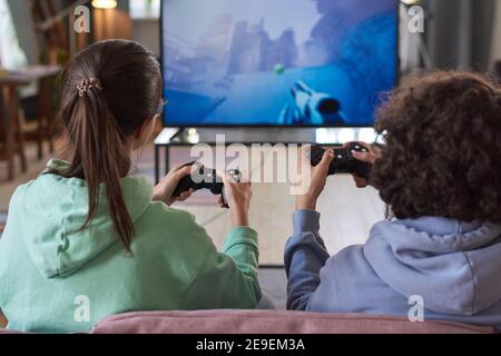 Vista posteriore di due amici che giocano a un videogioco in squadra Seduto sul divano di fronte alla grande TV Foto Stock