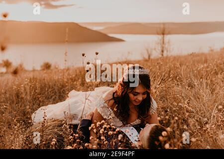 sposi in amore sposi sposi novelli in un vestito bianco e vestito stanno camminando che giacciono campo d'erba lungo in estate. l'uomo e la donna baciano. sposa e sposo hanno f Foto Stock