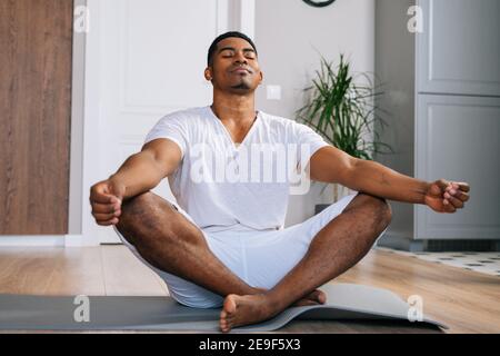 Uomo afro-americano rilassato con occhi chiusi meditando a casa seduta in posizione lotus sul tappetino. Foto Stock