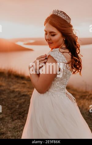 giovane sposa di brunette in abito da sposa bianco con corona sulla sua testa si erge su una scogliera sullo sfondo di Il fiume e le isole dopo il tramonto Foto Stock