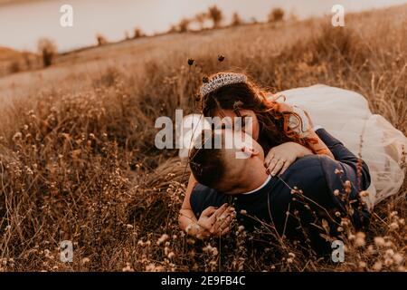 sposi in amore sposi sposi novelli in un vestito bianco e vestito stanno camminando che giacciono lungo campo d'erba in estate. l'uomo e la donna baciano. sposa e sposo hanno Foto Stock