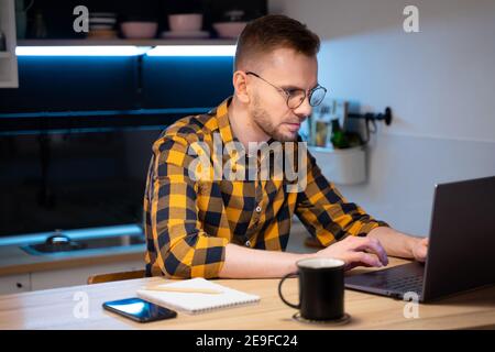 Ritratto del programmatore maschile in una camicia giallo-nero-grigio in una gabbia e occhiali utilizzando programmi codice computer in una casa. Lavora in modo professionale su un notebook Foto Stock