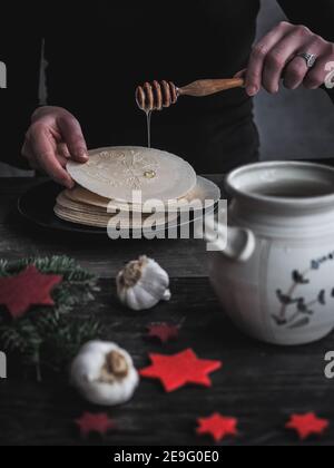 Mani femminili che versano il miele sui wafer della vigilia di Natale. Pentola di miele e bulbi di aglio su tavolo di legno scuro. Buio e moody. Foto Stock