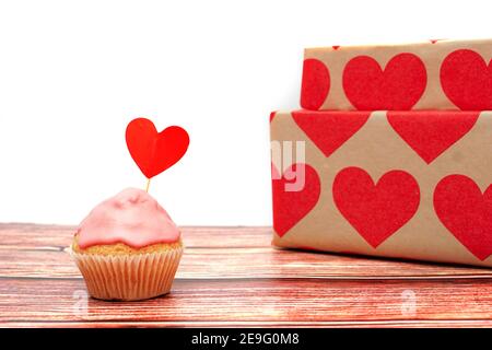 Due presenta avvolti in cuori avvolgenti su una superficie di legno e uno sfondo vuoto con spazio di copia, con una cupcake glassante rosa con un bastone cardiaco Foto Stock