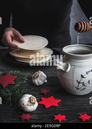 Mani femminili che versano il miele sui wafer della vigilia di Natale. Pentola di miele e bulbi di aglio su tavolo di legno scuro. Buio e moody. Foto Stock