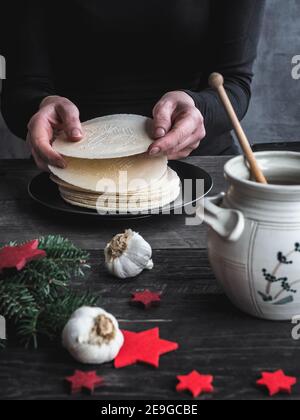 Mani femminili che tengono i wafer della vigilia di Natale. Pentola di miele, bulbi di aglio e decorazione di Natale su tavola di legno. Scuro e moddy. Foto Stock