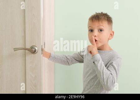 Carino ragazzo che mostra il gesto del silenzio e l'apertura della porta dentro camera Foto Stock